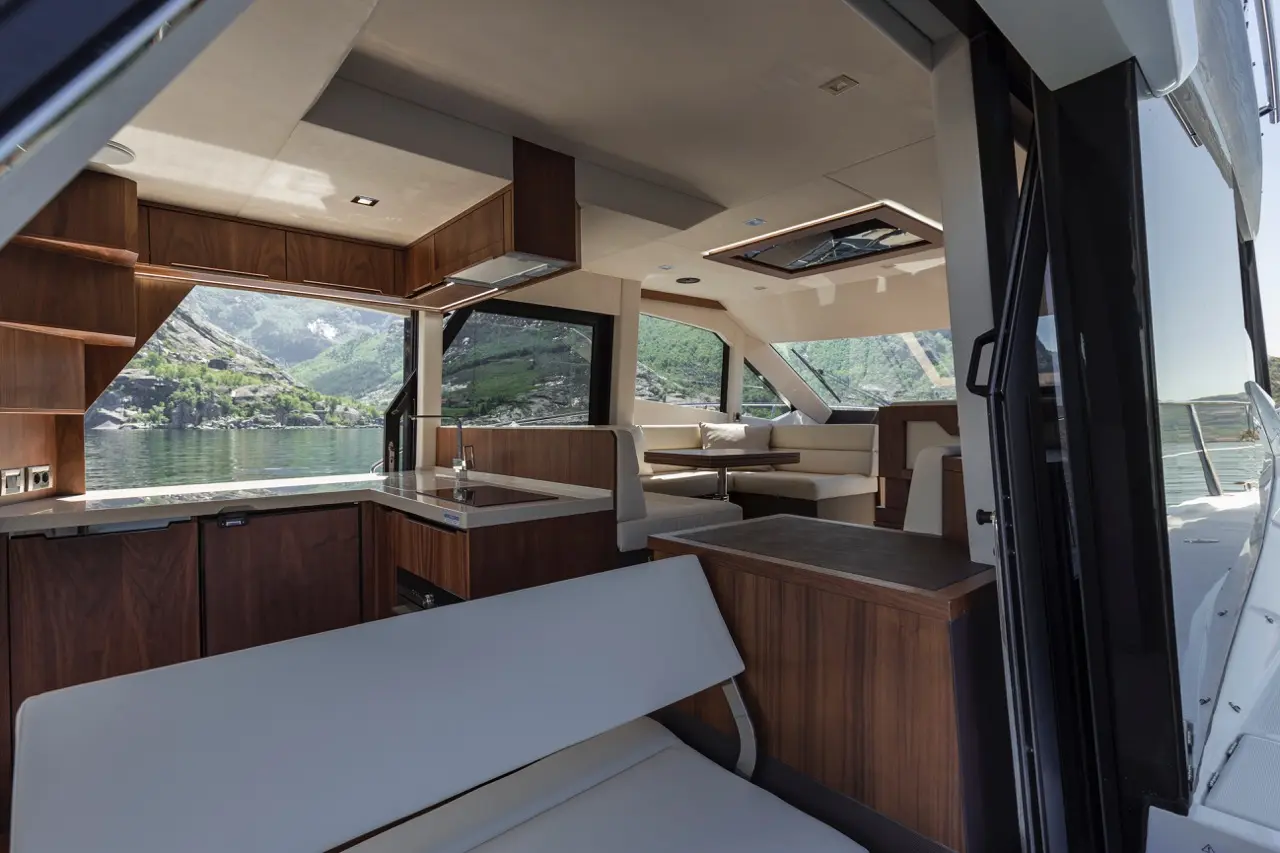 Modern yacht interior showcasing a dining area with scenic views of green hills and water, featuring wood and white decor.