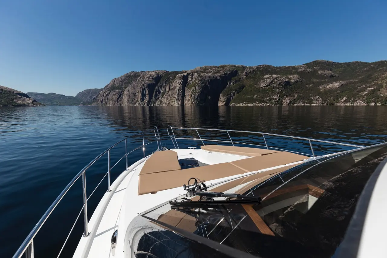 A boat's bow with lounge seats, reflecting calm water and steep, rocky cliffs under a clear blue sky.