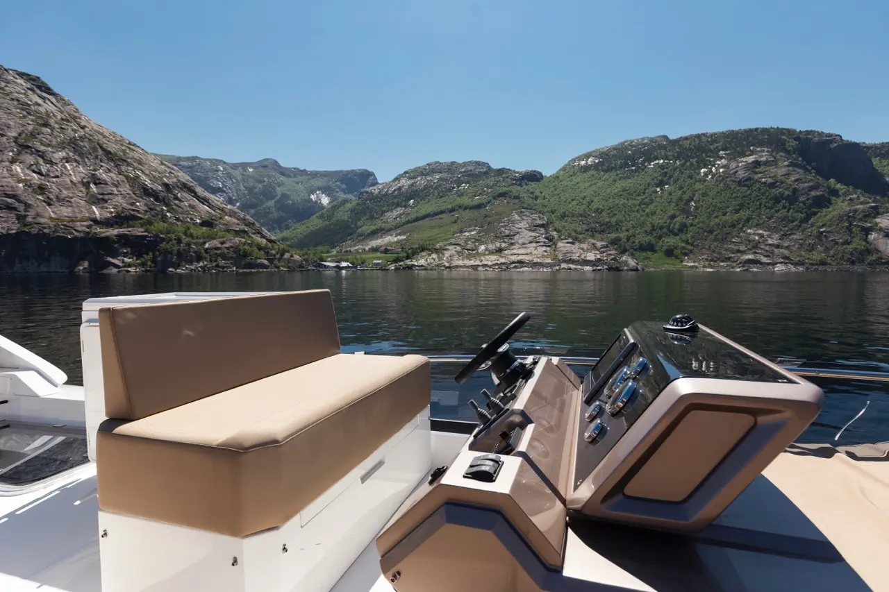 A stylish boat's helm area features a beige seat and modern controls, with lush green mountains reflected in calm waters.