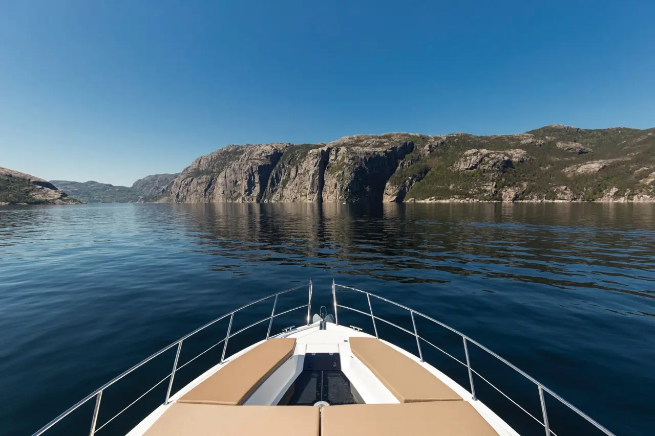 Bow of a boat on calm waters, with steep cliffs and greenery reflecting in the water under a clear blue sky.