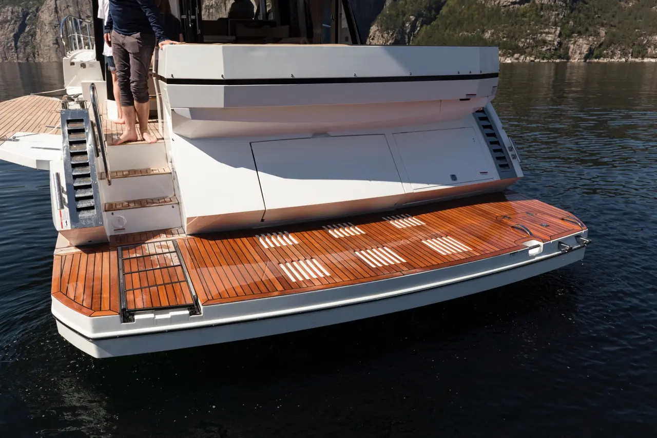 A luxury yacht's deck with wooden flooring, stairs, and a scenic backdrop of rocky cliffs and clear water.
