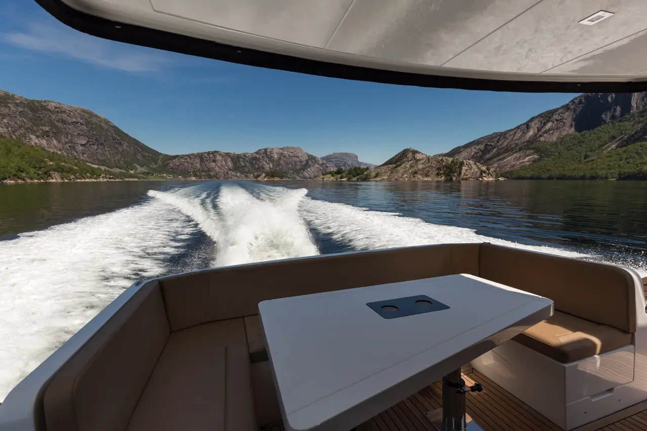 Scenic view from a boat's interior, showcasing a table and mountains reflecting in calm waters under a clear blue sky.