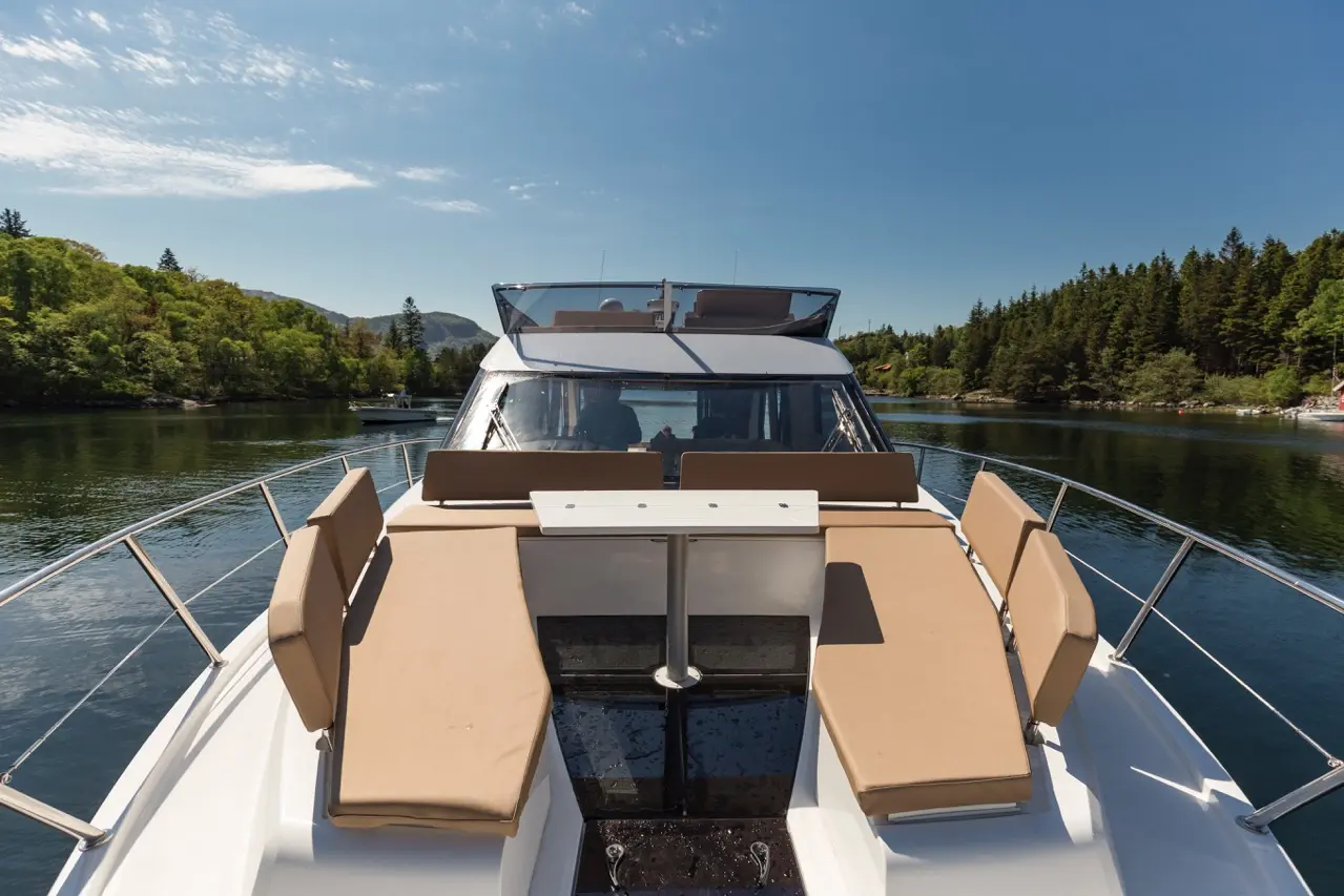 A luxury boat's back deck featuring tan seating, a central table, and serene waters surrounded by lush greenery and hills.