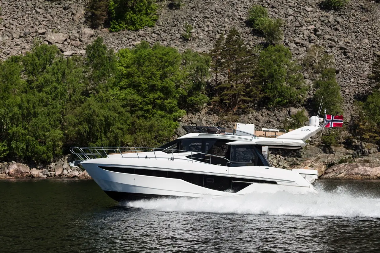A white luxury motorboat cruises through calm waters near rocky shores, featuring a Norwegian flag flying from its stern.