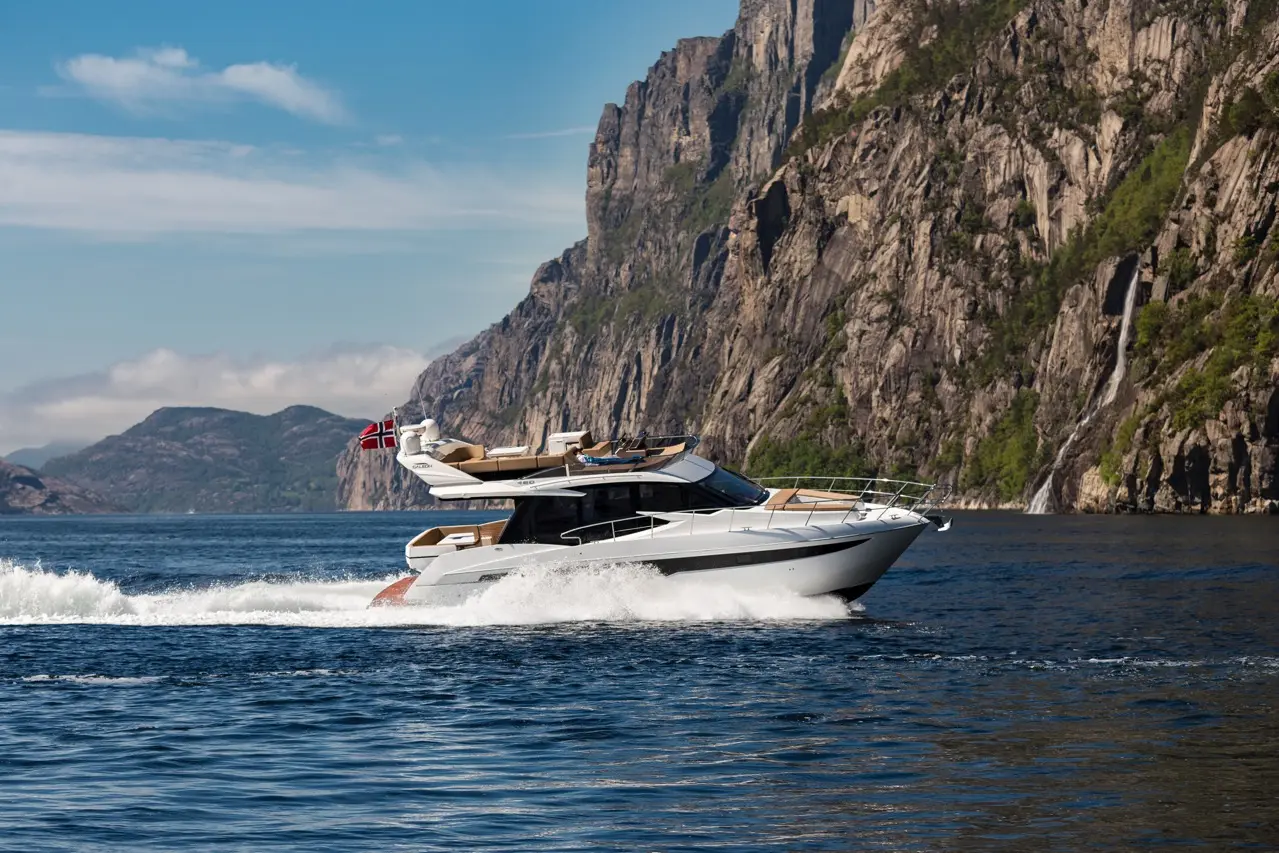 A yacht with a Norwegian flag speeds through calm waters, surrounded by dramatic cliffs and lush greenery in the background.