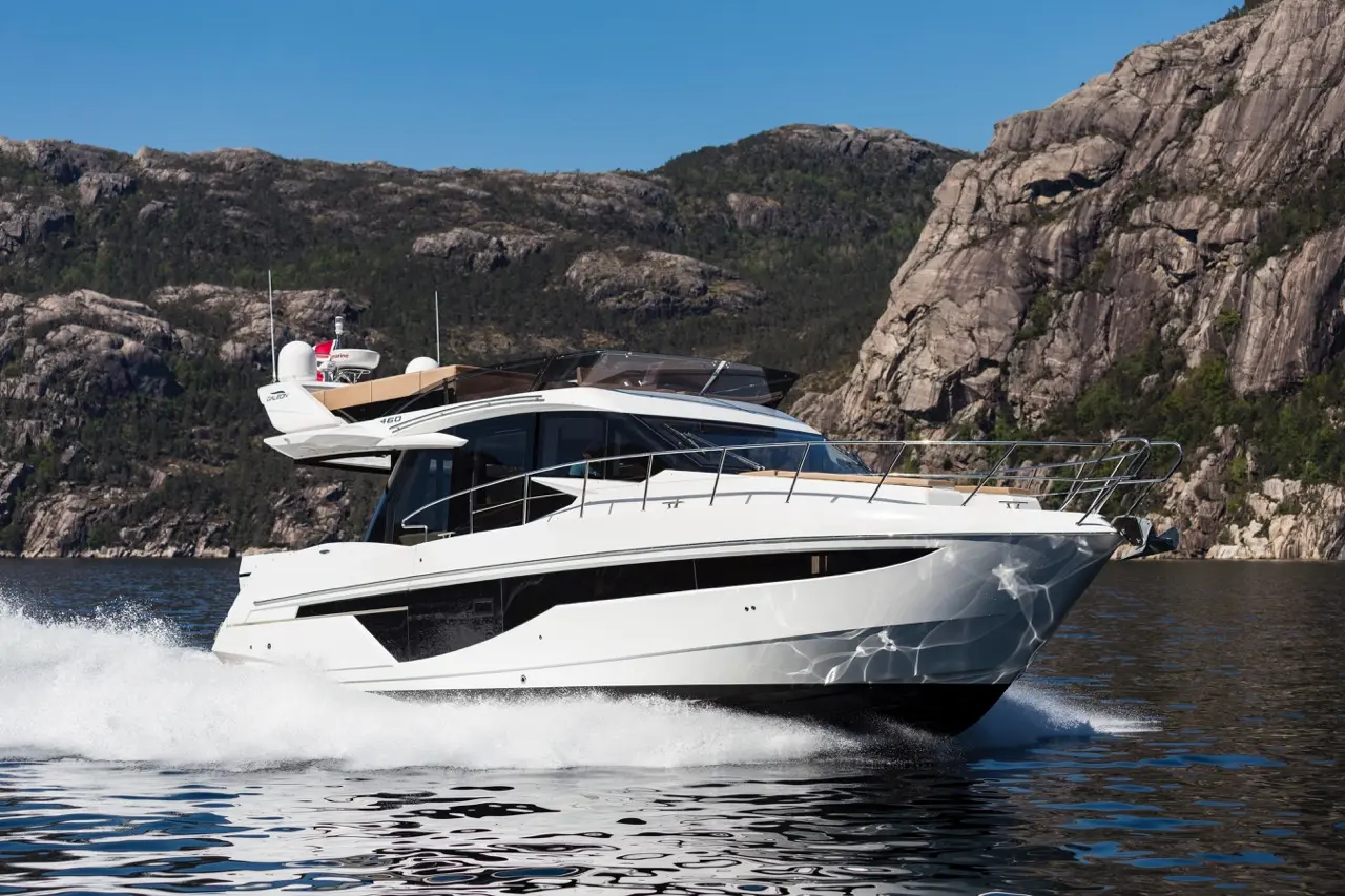 A sleek white motor yacht glides through calm waters, with rugged mountains and greenery in the background under a clear blue sky.