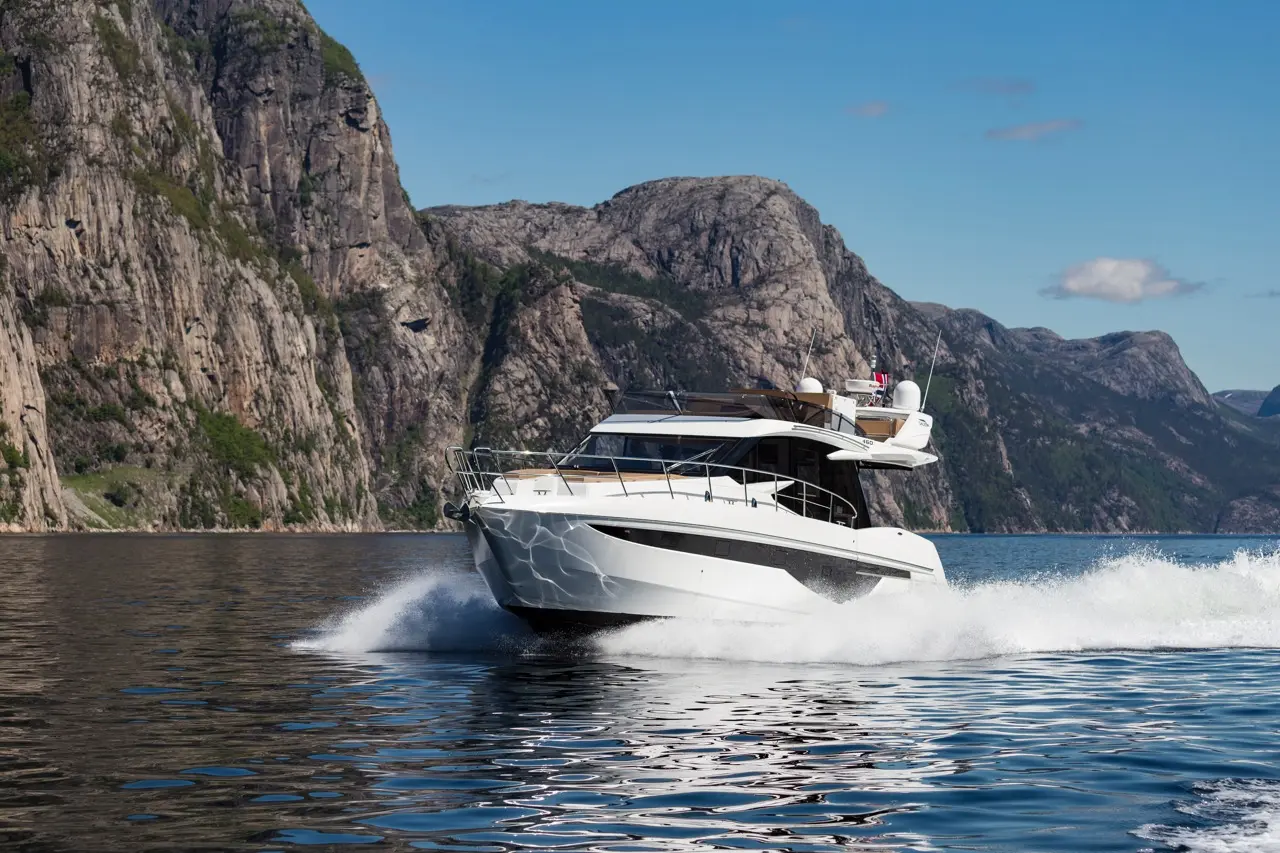 A sleek white motorboat speeds across a calm fjord, surrounded by rugged cliffs and blue skies.