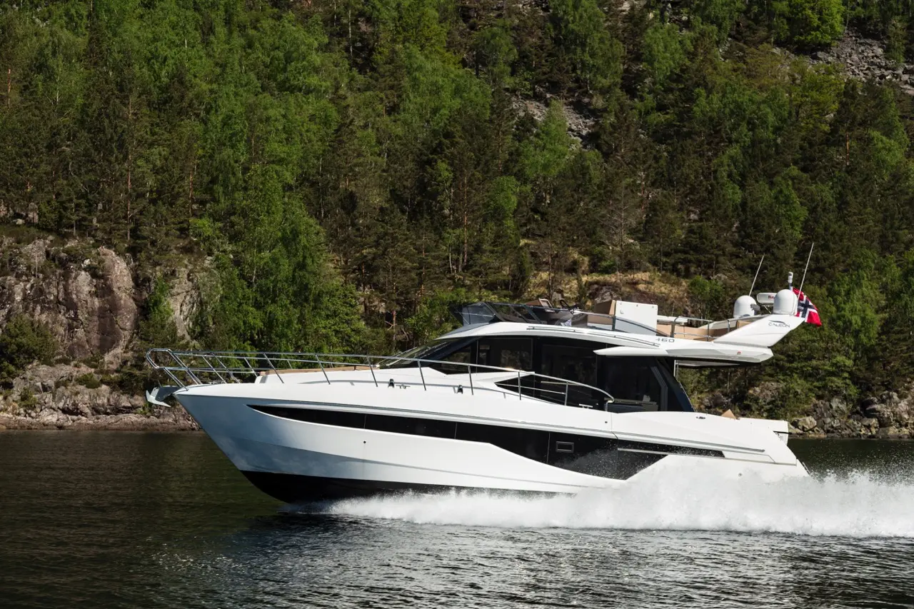 A sleek white yacht speeds across a calm waterway, surrounded by lush green trees and rocky shores.