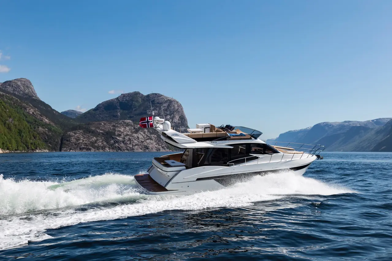 A sleek motor yacht with a Norwegian flag speeds across sparkling blue water, surrounded by majestic mountains and greenery.