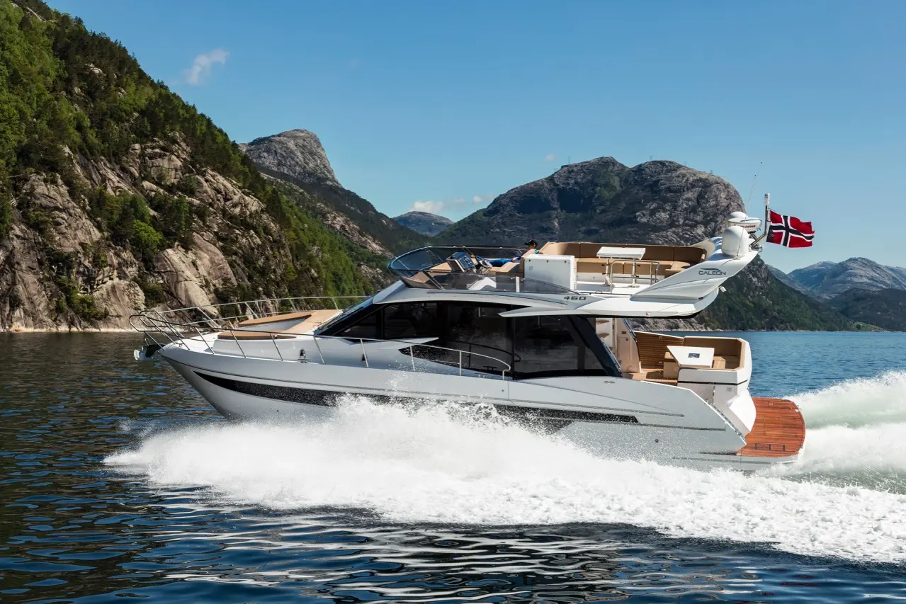 A modern motor yacht cruising through clear waters near rocky hills, with a Norwegian flag waving in the background.