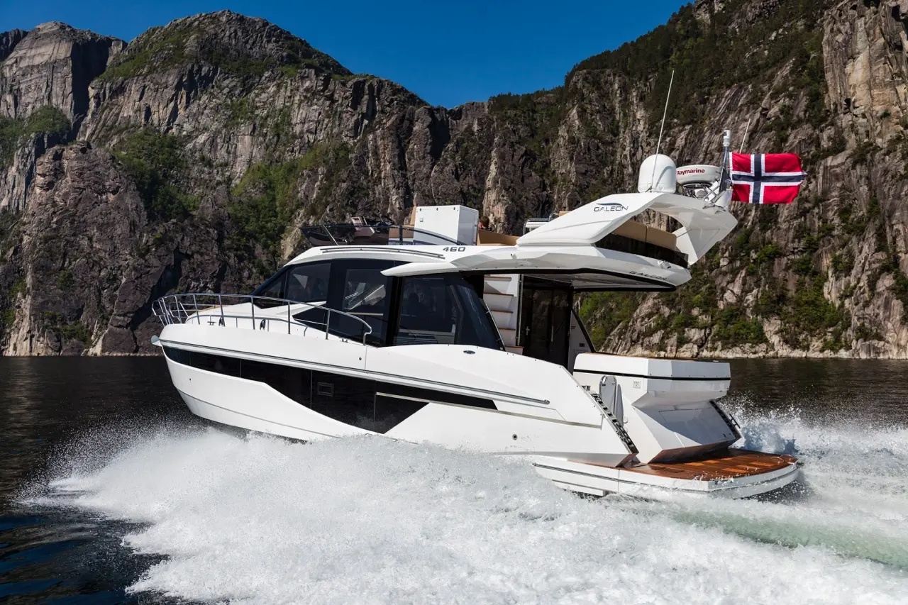 A sleek white motorboat speeds through water with a Norwegian flag, surrounded by rocky cliffs and clear blue skies.