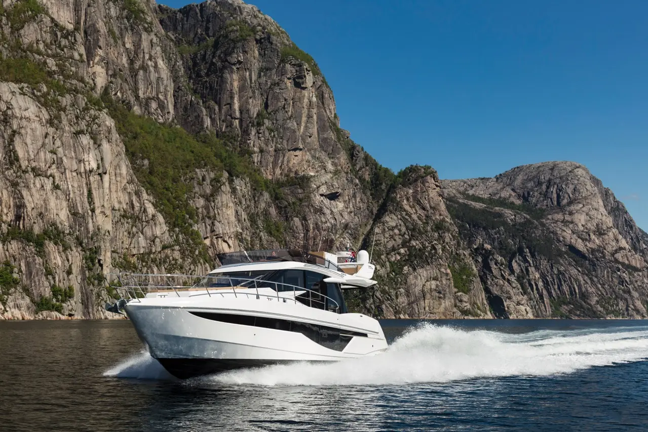 A luxurious motor yacht glides over calm waters, set against majestic rocky cliffs under a clear blue sky.
