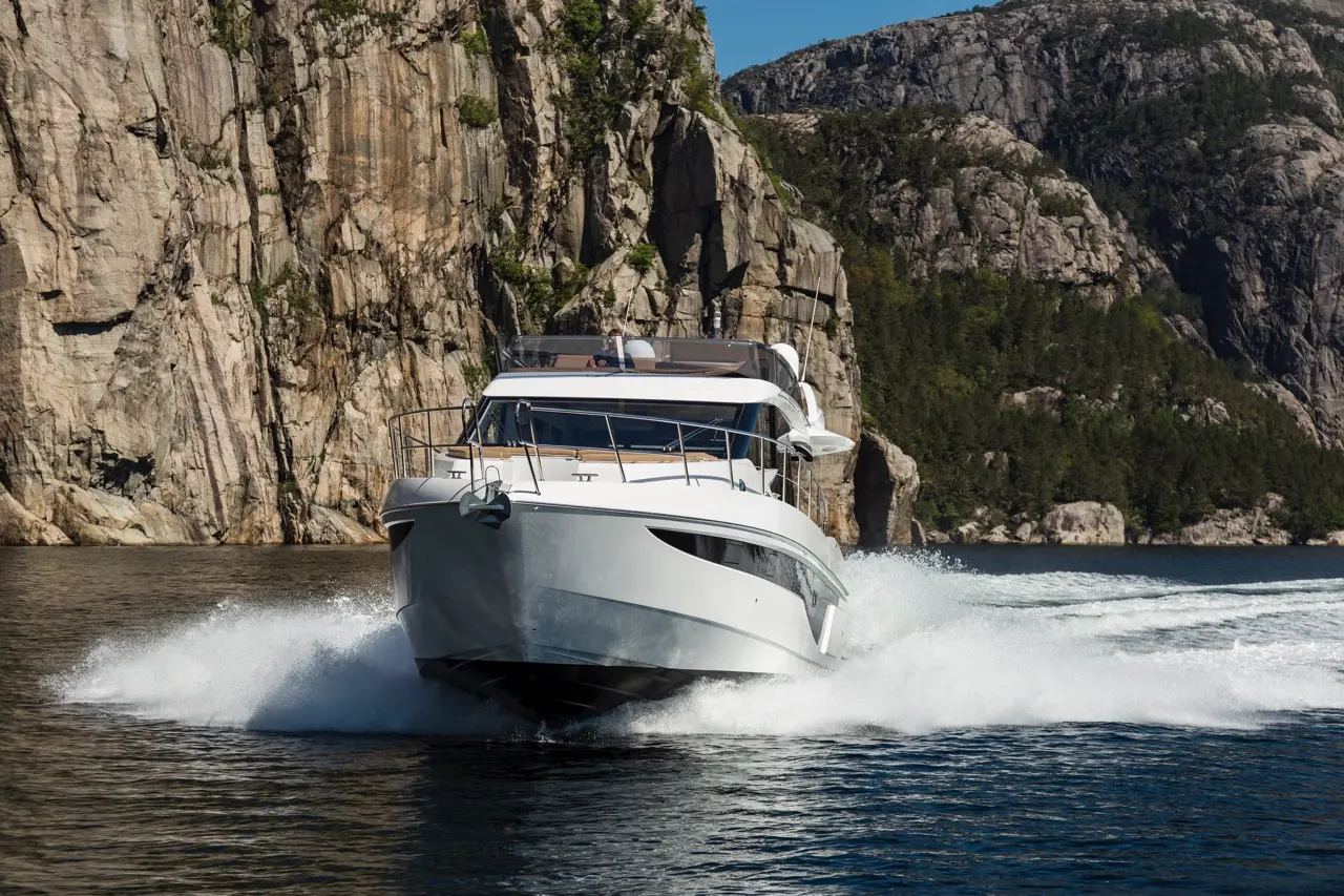 A white motorboat sails through clear waters, creating sprays, with rocky cliffs and greenery in the background.