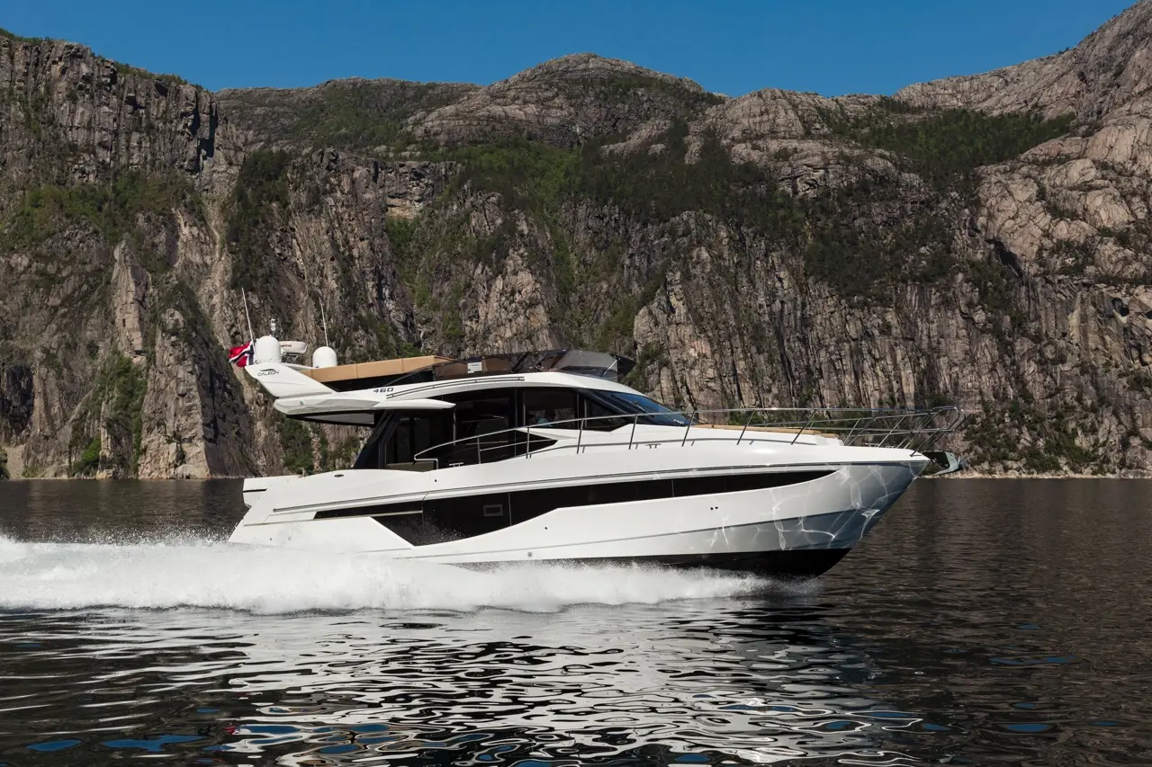 A sleek white yacht cruises through calm waters, flanked by towering rocky cliffs and clear blue skies.