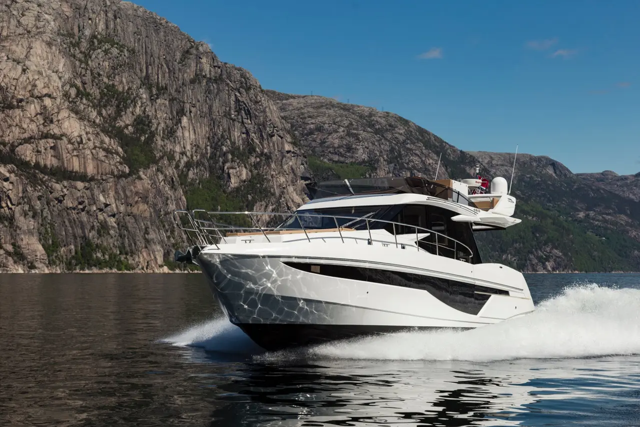 A sleek white motor yacht glides over calm water, framed by rugged cliffs under a clear blue sky.