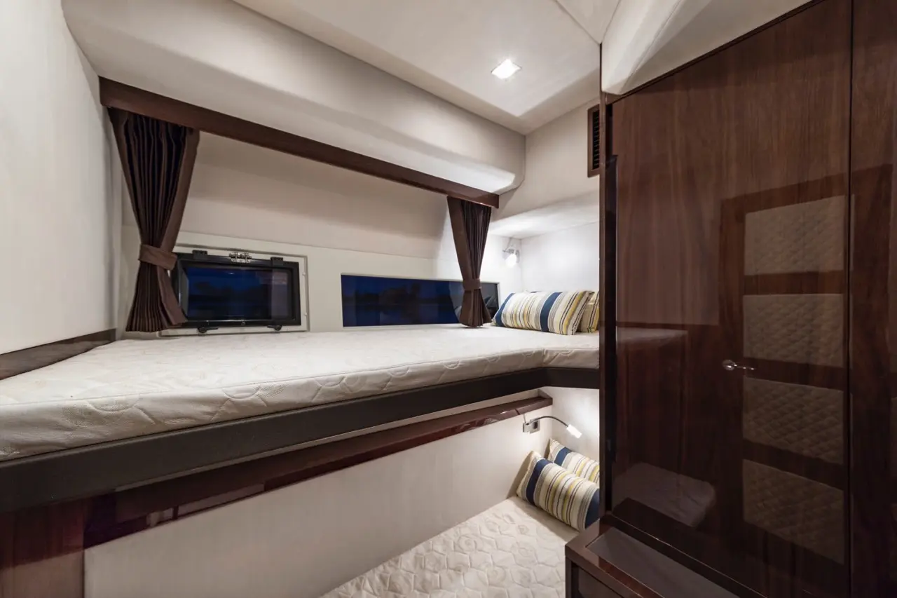 Modern boat cabin featuring two beds, decorative cushions, a television, and a wooden wardrobe, softly lit for a cozy atmosphere.