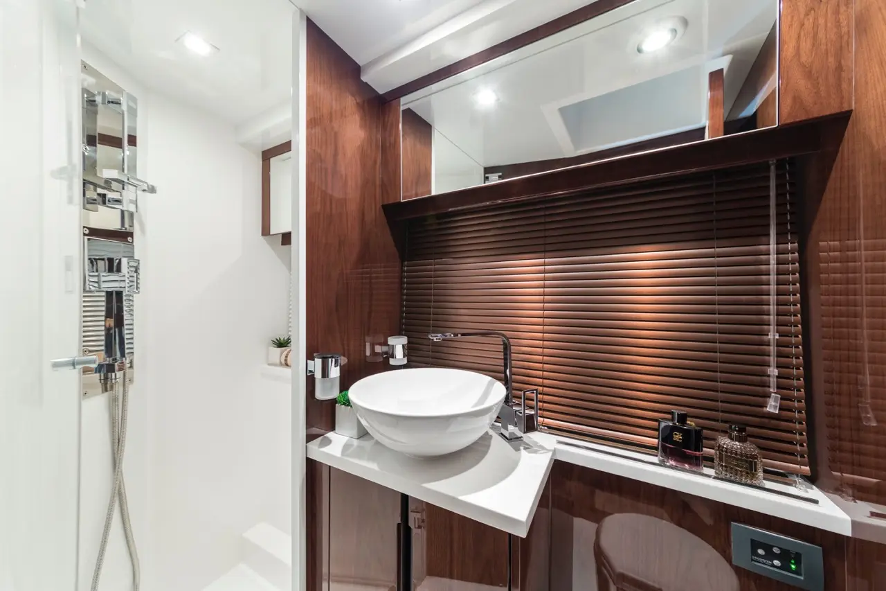 Modern bathroom with a round white basin, wooden accents, blinds, and toiletries on a countertop. Brightly lit and stylish.