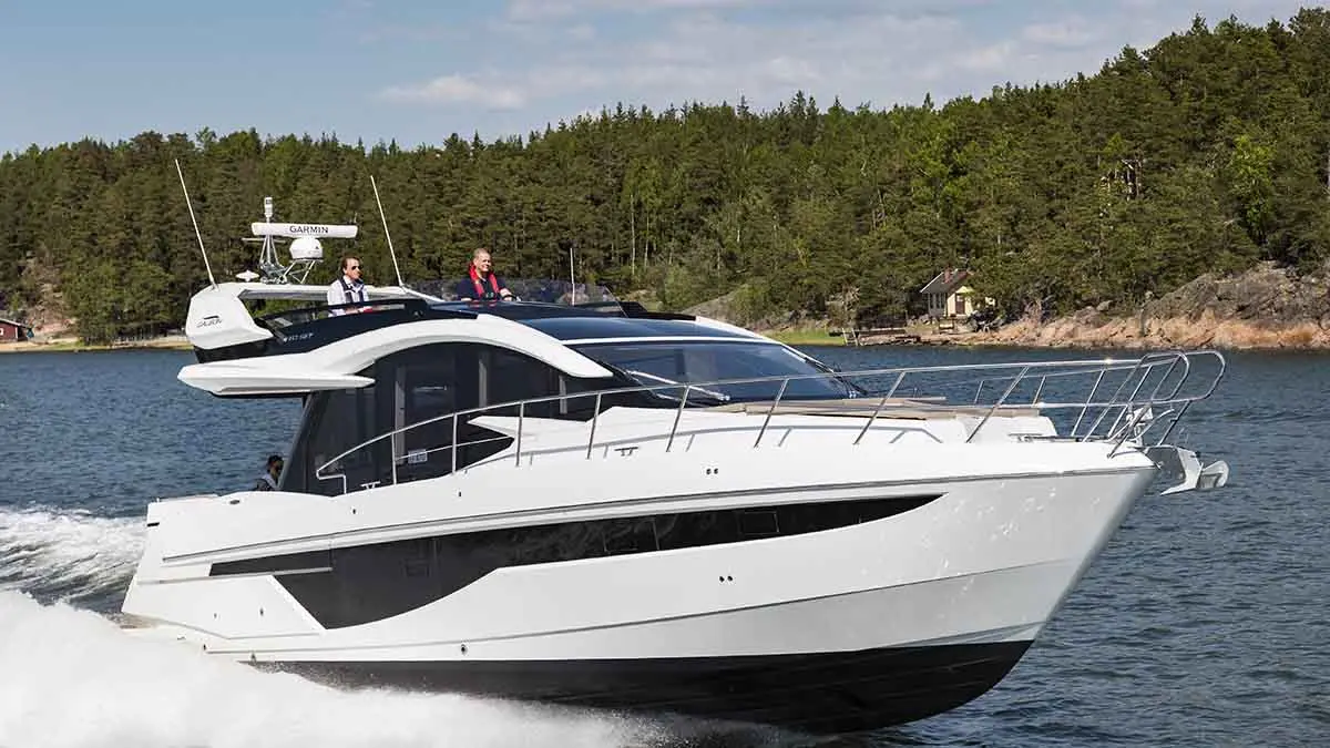 A modern white motorboat cruises through water with two people onboard, surrounded by a forested landscape.