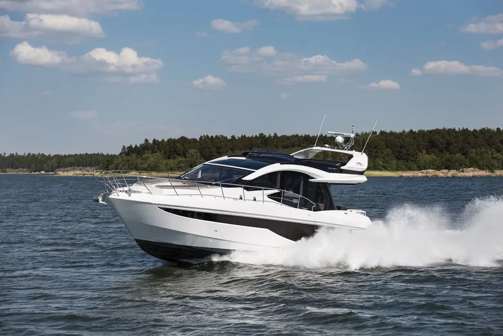 A sleek white motorboat speeds through blue water, surrounded by trees and under a partly cloudy sky.