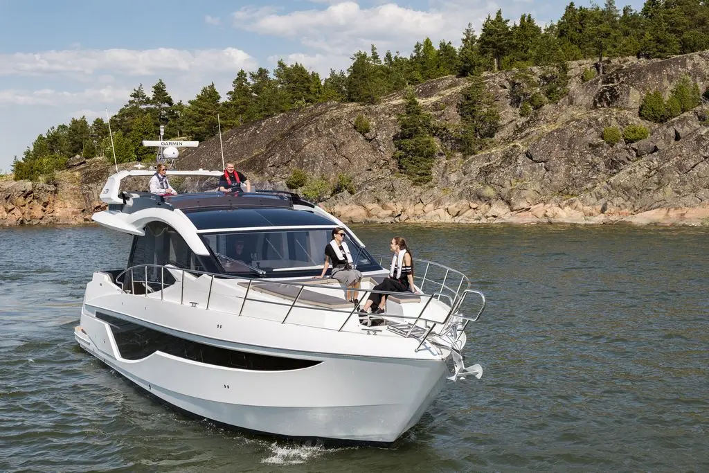 A sleek white motor yacht navigates through calm waters, with four people relaxing onboard against a rocky shoreline.