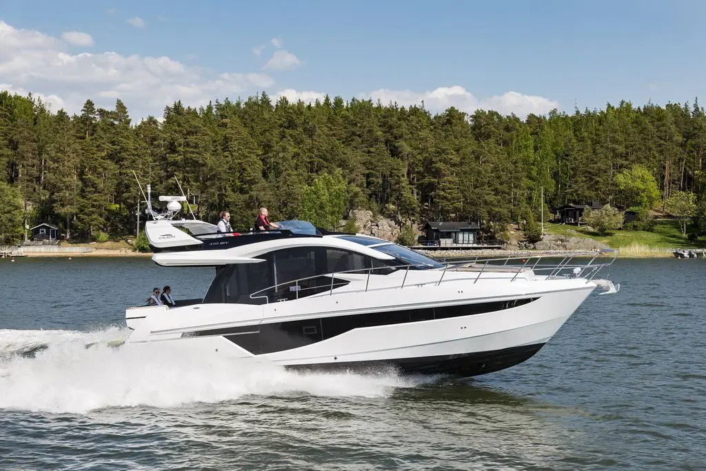 A sleek motor yacht speeds through calm waters, with people on board enjoying the sunny scenery of lush greenery.