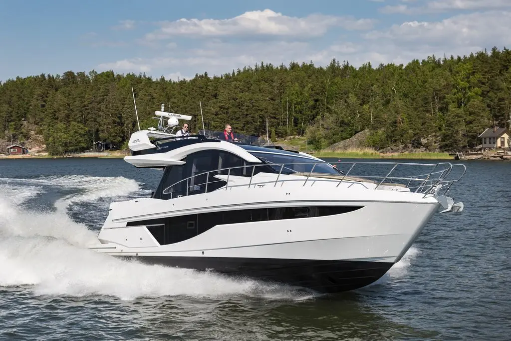 A sleek white motorboat navigates through water with two people aboard, surrounded by lush green trees and a serene shoreline.