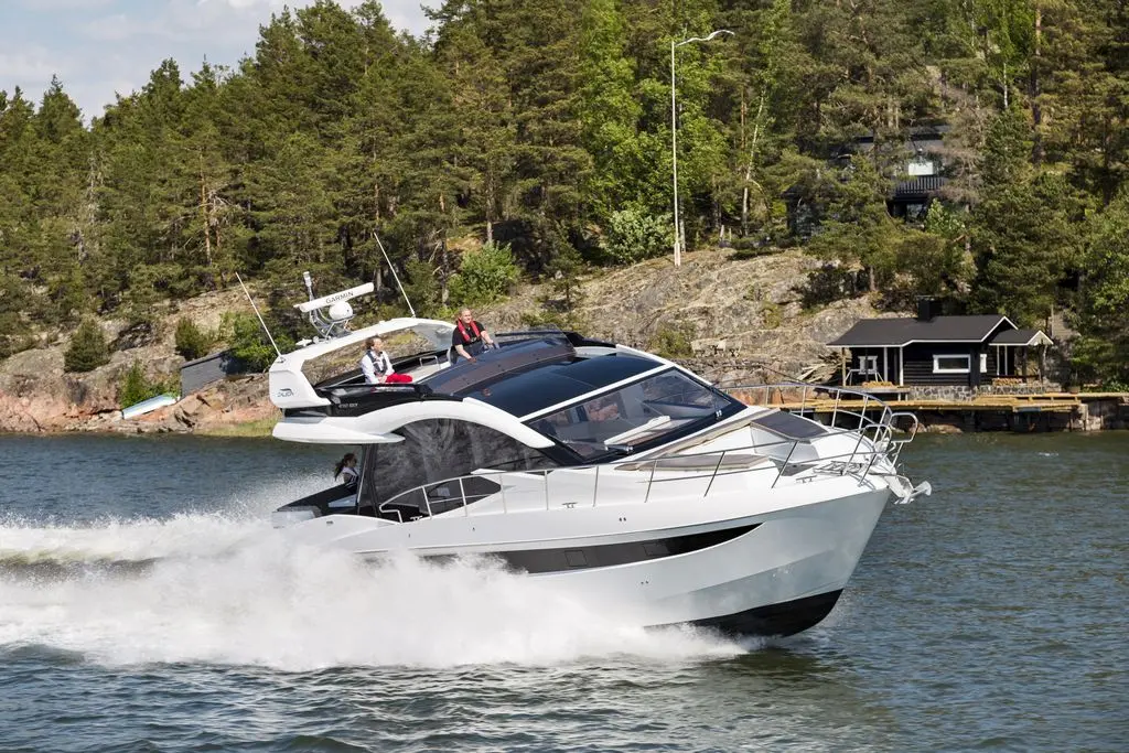 A sleek white motorboat speeds through water with two people onboard, surrounded by green trees and a lakeside cottage.