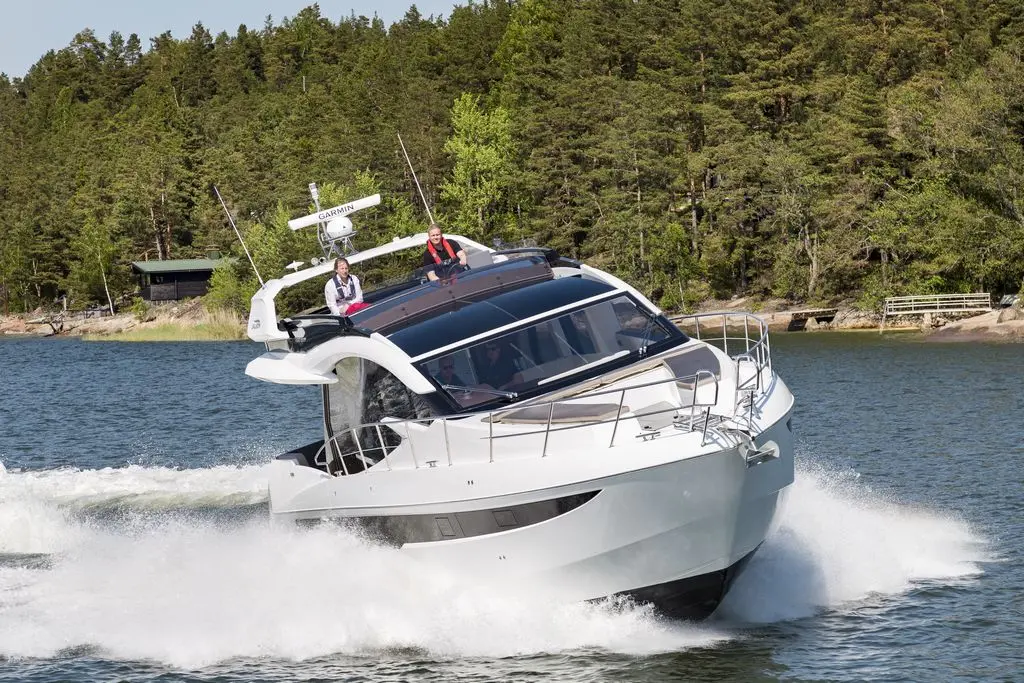 A sleek white yacht speeds across a calm lake, creating a spray of water, with two people enjoying the ride on deck.