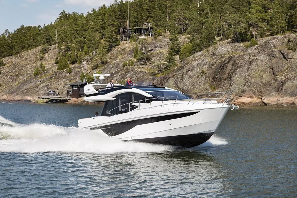 A sleek white motorboat speeds through calm waters near rocky shores, surrounded by green trees and a wooden cabin.