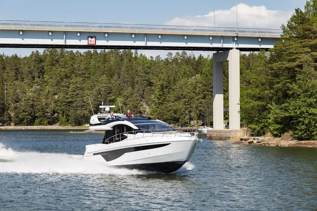 A sleek white motorboat speeds through a calm river beneath a bridge, surrounded by lush green trees and clear blue skies.