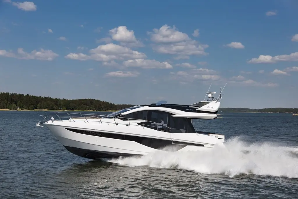 A sleek white motor yacht cruising on a calm lake under a blue sky with fluffy clouds and green forests in the background.