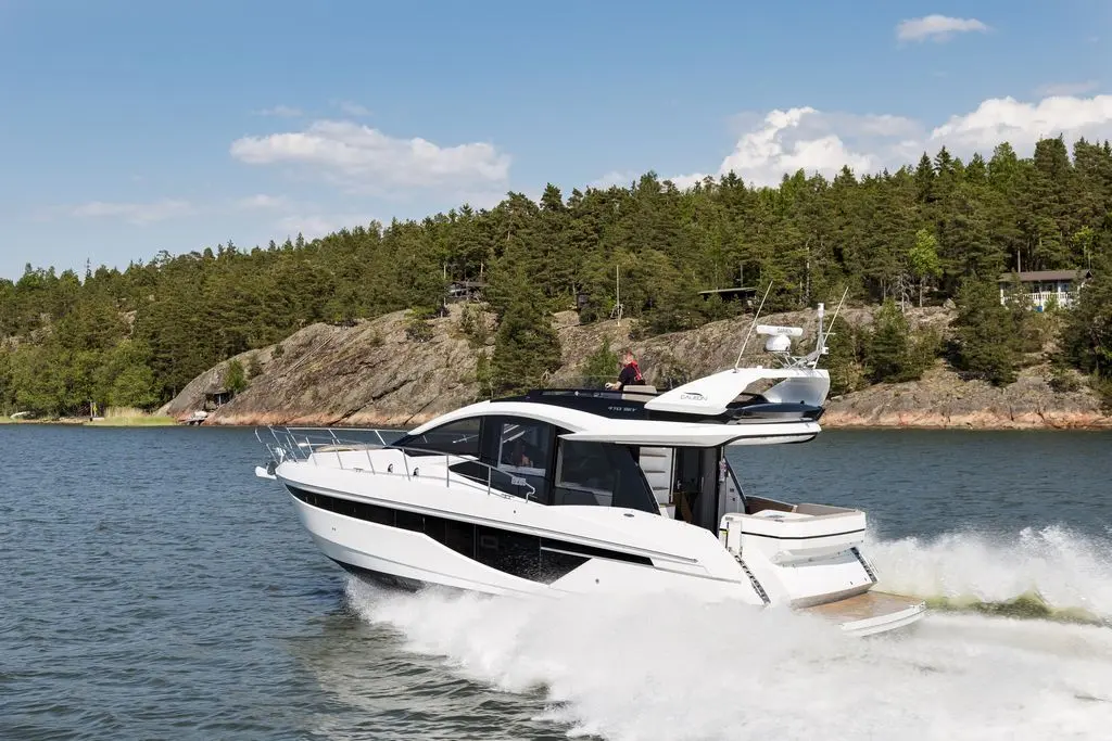 A stylish motor yacht speeds across calm waters, surrounded by green forests and rocky shores under a clear blue sky.