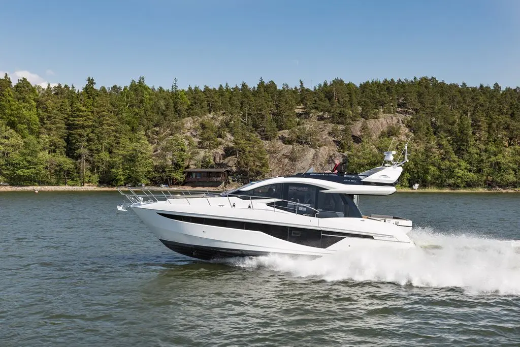 A sleek white motorboat speeding across calm water, surrounded by green trees and rocky shore in a sunny setting.