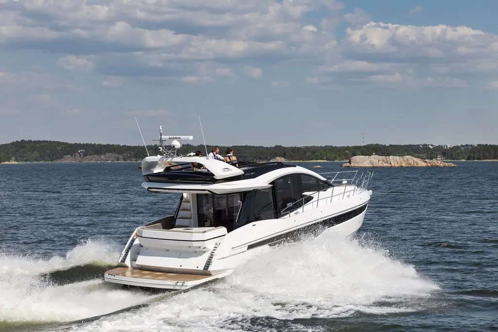 A sleek motorboat with passengers cruises through calm waters, leaving a wake against a scenic backdrop of trees and rocks.