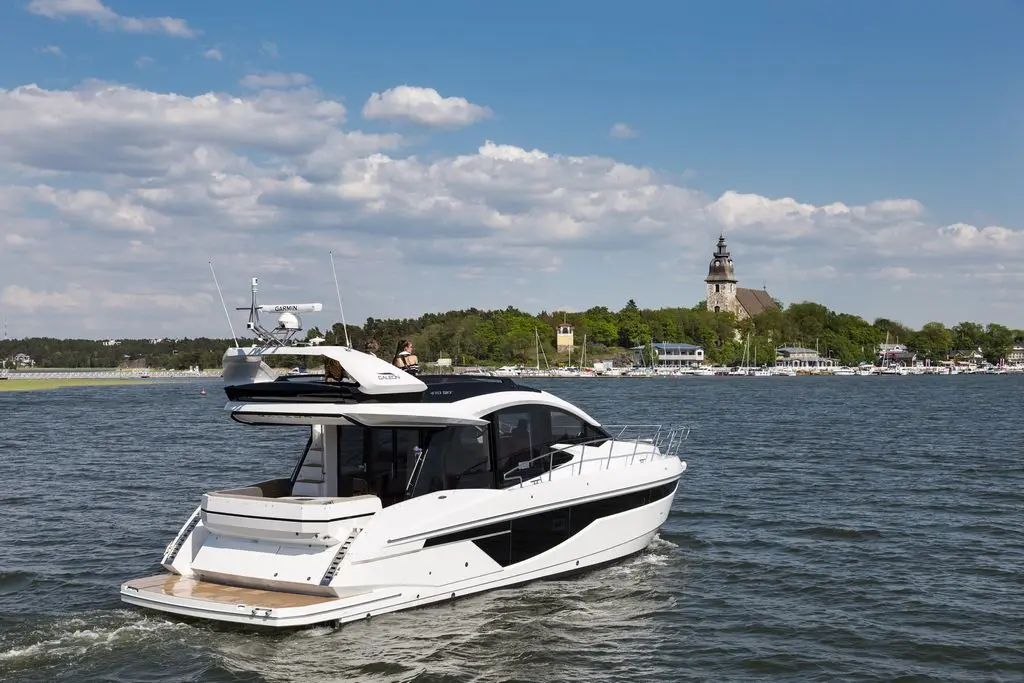 A sleek white yacht navigates a calm waterway, with a historic church and green trees in the background under a blue sky.