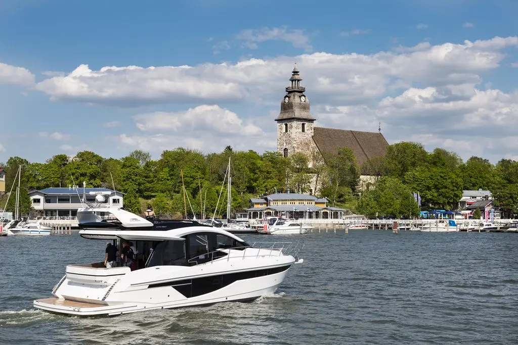 A white motorboat navigates a scenic waterfront with lush greenery and a historic church in the background under blue skies.