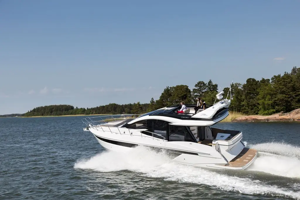 A sleek white yacht speeds across calm water, surrounded by green trees and rocky shore, with people enjoying the ride.