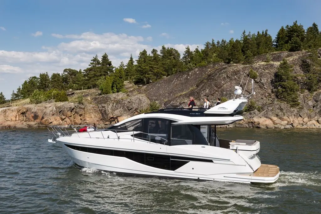A sleek white motor yacht glides through water near a rocky shore, surrounded by lush green trees under a clear blue sky.