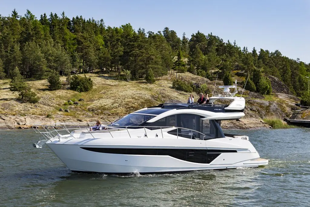 A white motor yacht glides across calm water, surrounded by lush green trees and rocky shorelines, with people onboard enjoying the view.