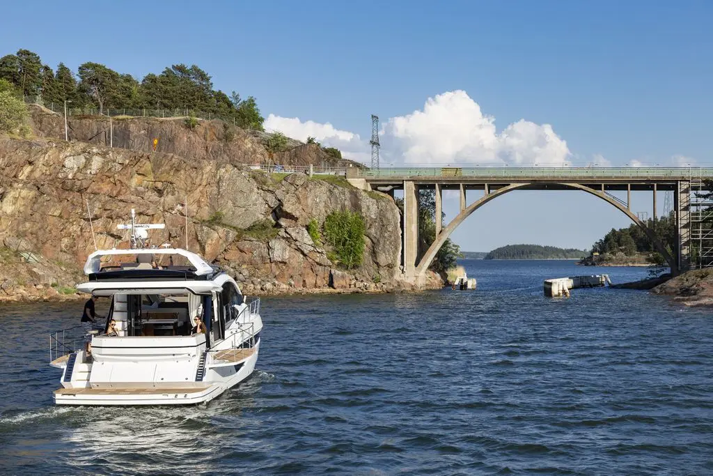A luxury boat navigates a calm waterway beneath a stone bridge, surrounded by greenery and rocky landscapes.