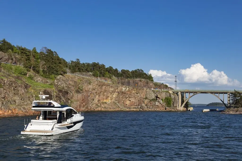 A motorboat navigates a rocky shoreline under a clear blue sky, with a bridge spanning across the scene.