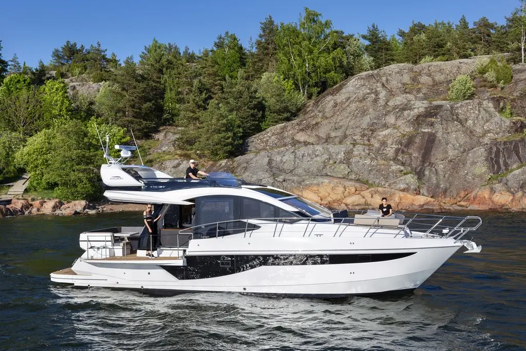 Luxury yacht cruising on calm waters with three people aboard, surrounded by lush greenery and rocky shoreline.