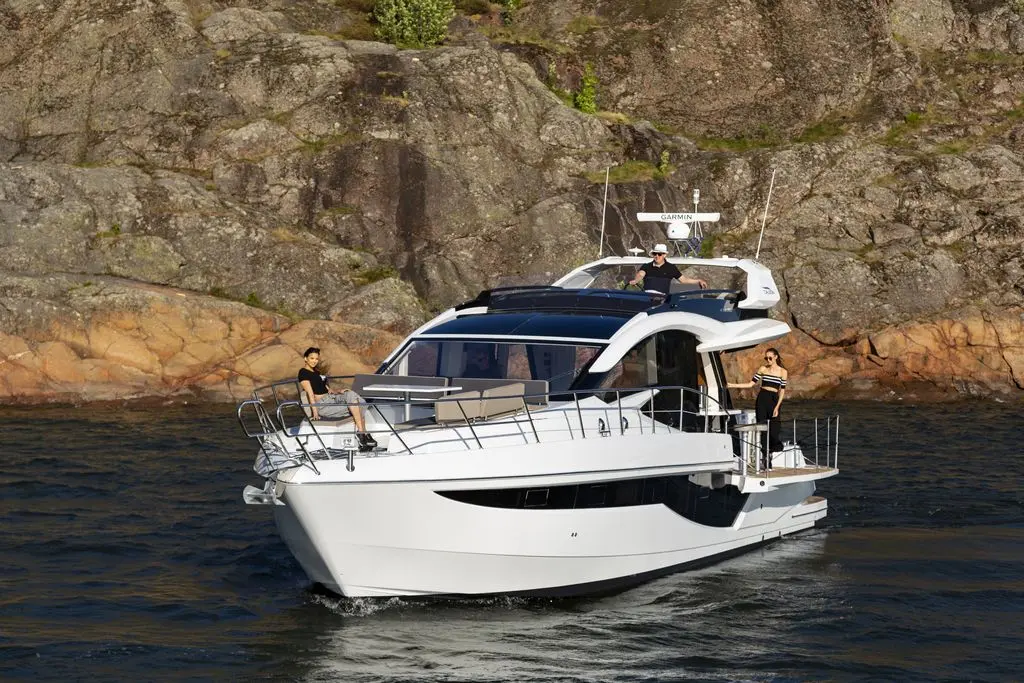 A luxury yacht sails on calm waters, with three people casually enjoying the sun against a rocky shoreline backdrop.