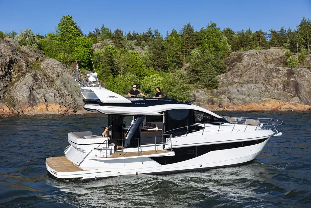 A modern yacht cruising in a serene waterway, surrounded by lush greenery and rocky shores, with people on board enjoying the view.