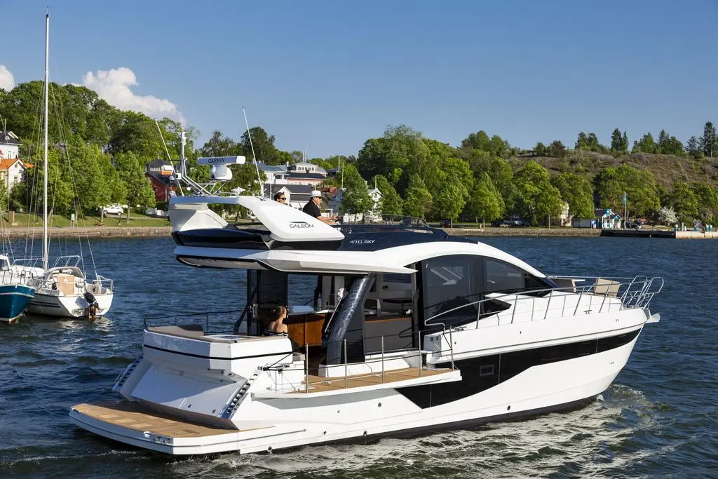 A sleek white motor yacht with a black stripe navigates a calm waterway surrounded by greenery and distant buildings.