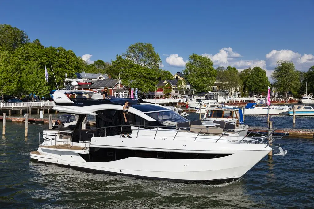 A sleek white luxury yacht sails in a calm harbor, surrounded by green trees and various boats, under a clear blue sky.