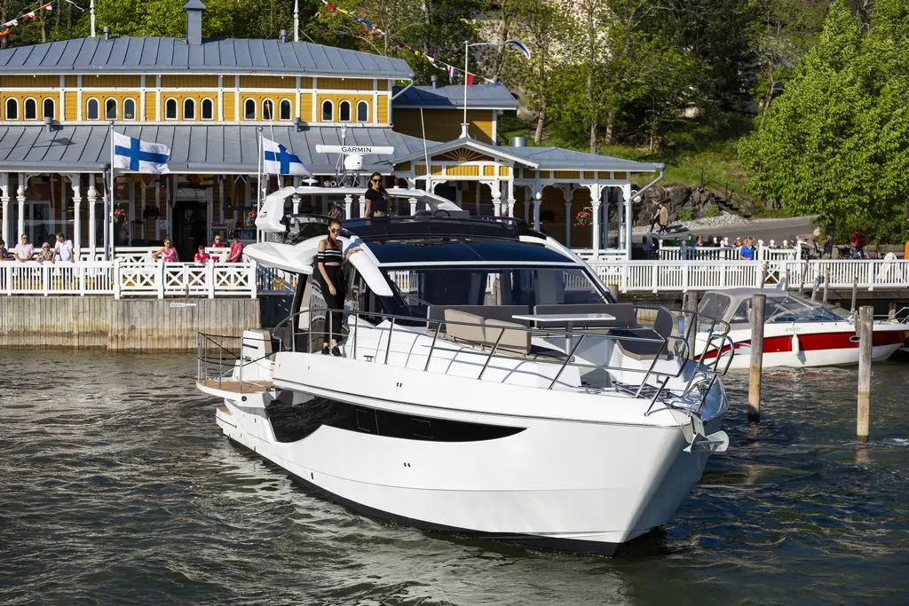 A sleek white yacht approaches a waterfront, with people visible on the dock and Finnish flags flying.