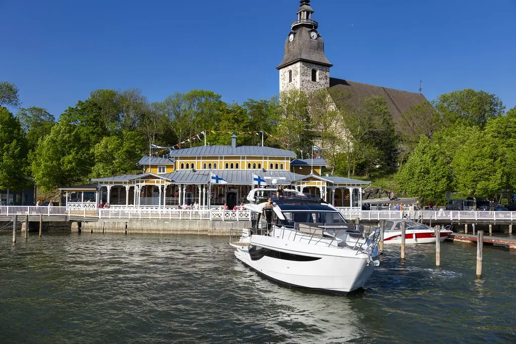 A white boat approaches a lakeside restaurant with colorful trim, surrounded by greenery and a church tower in the background.