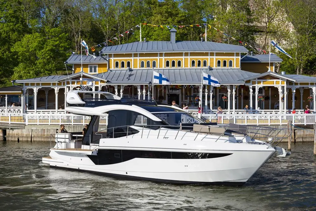 A luxury yacht navigates a lake near a vibrant yellow building adorned with Finnish flags and greenery.