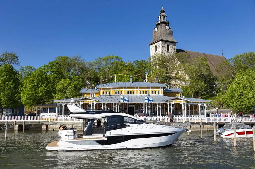 A modern yacht moored near a charming lakeside pavilion and a historic church, surrounded by lush greenery and blue skies.